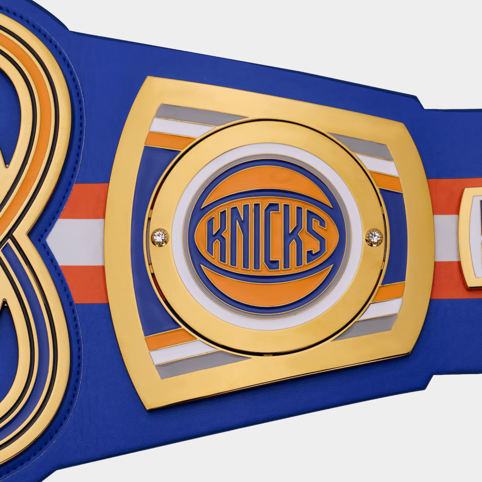Close-up of a New York Knicks championship-style belt or decorative strap with a large gold and blue central plaque featuring the Knicks logo, blue leather-like base, and orange and white striping.