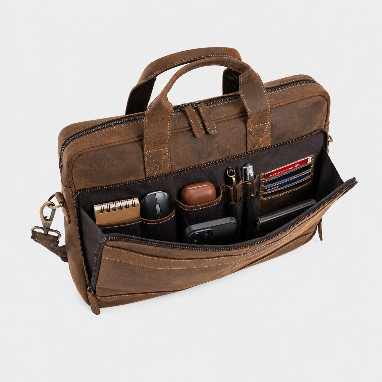 A brown leather briefcase shown open, revealing multiple interior compartments filled with office accessories such as a notebook, pens, cards, and a phone. The bag has dual top handles, a shoulder strap, and a zip-around closure, photographed on a clean light gray background.
