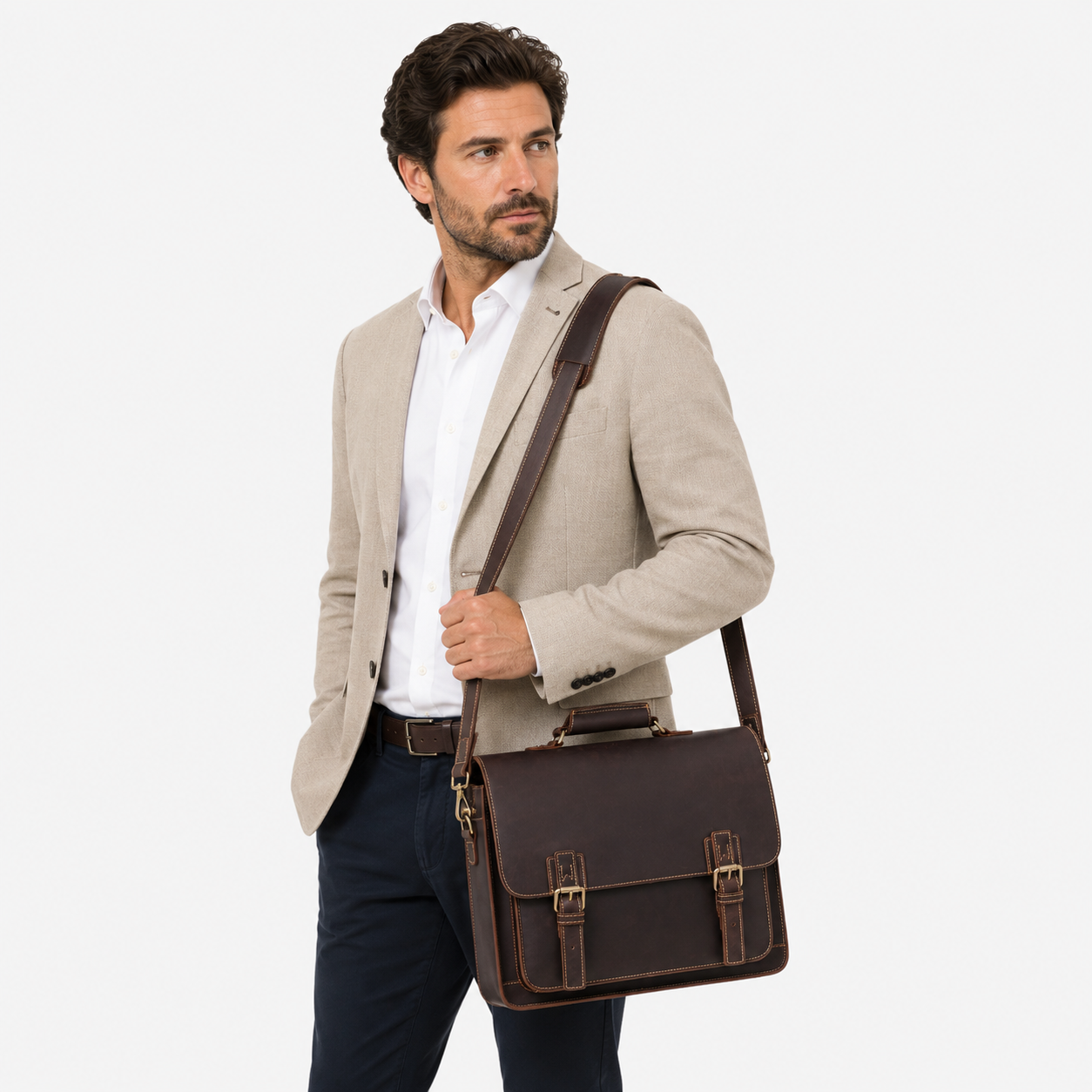 A man wearing a light beige blazer, white shirt, and navy trousers carries a dark brown leather satchel-style briefcase bag with a shoulder strap against a plain light gray studio background.