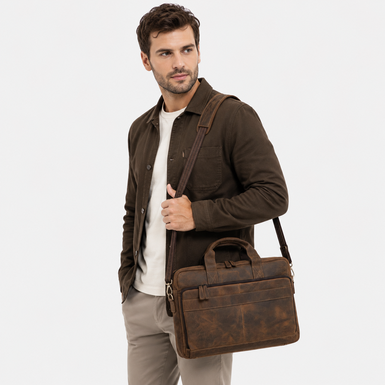 A man wearing a brown overshirt, white t-shirt, and beige trousers carries a brown distressed leather briefcase-style shoulder bag against a light gray studio background.