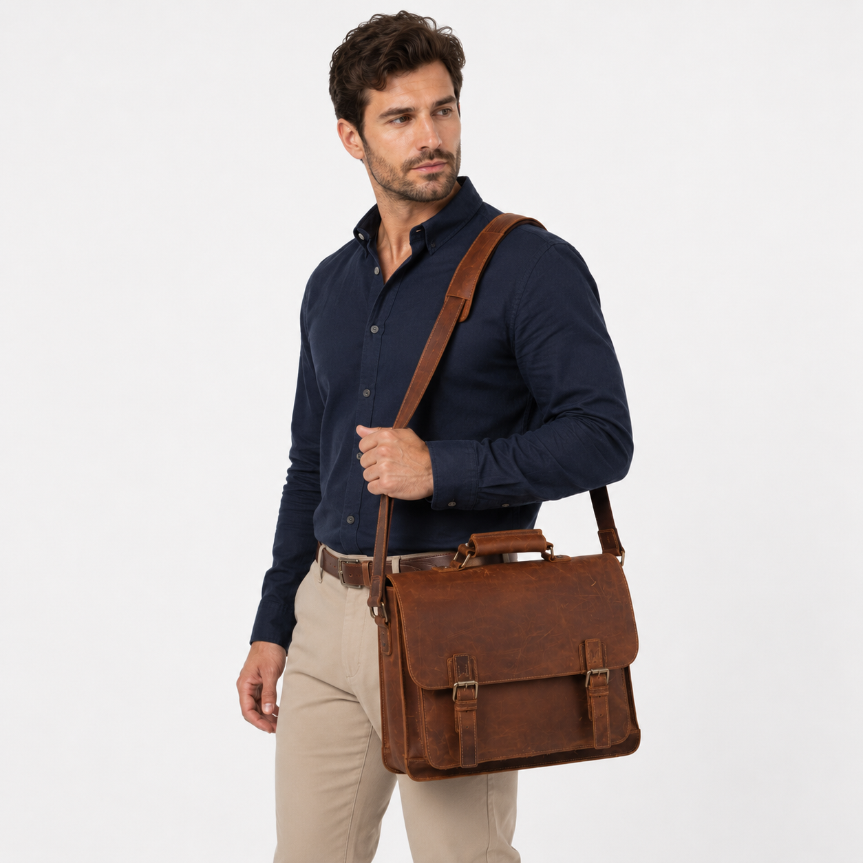 A man wearing a navy blue button-down shirt and beige trousers carries a brown leather briefcase-style shoulder bag against a plain light gray studio background.