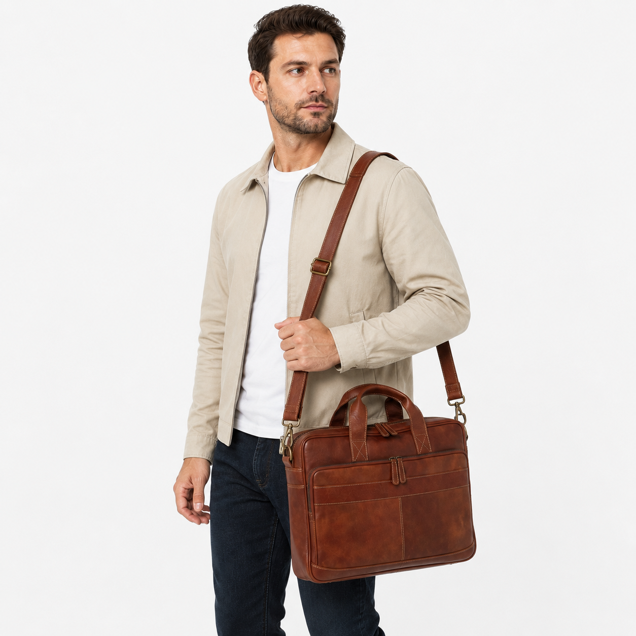 A man wearing a beige jacket, white t-shirt, and dark jeans carries a brown leather briefcase-style shoulder bag with a top handle and adjustable strap against a plain light gray studio background.