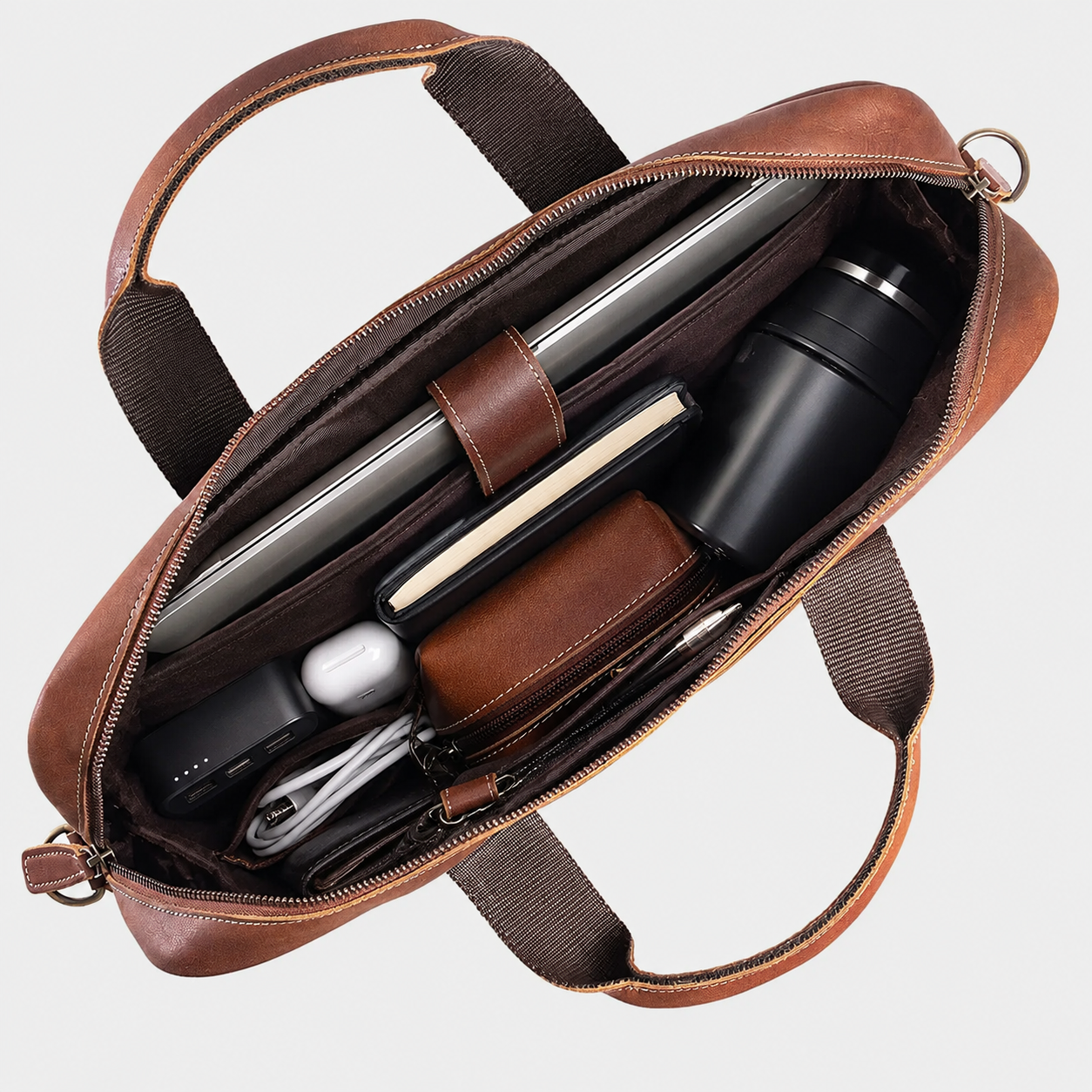 An open brown leather laptop bag shown from above, revealing organized interior compartments with a laptop, notebook, wallet, bottle, earbud case, and cables, on a light gray background.