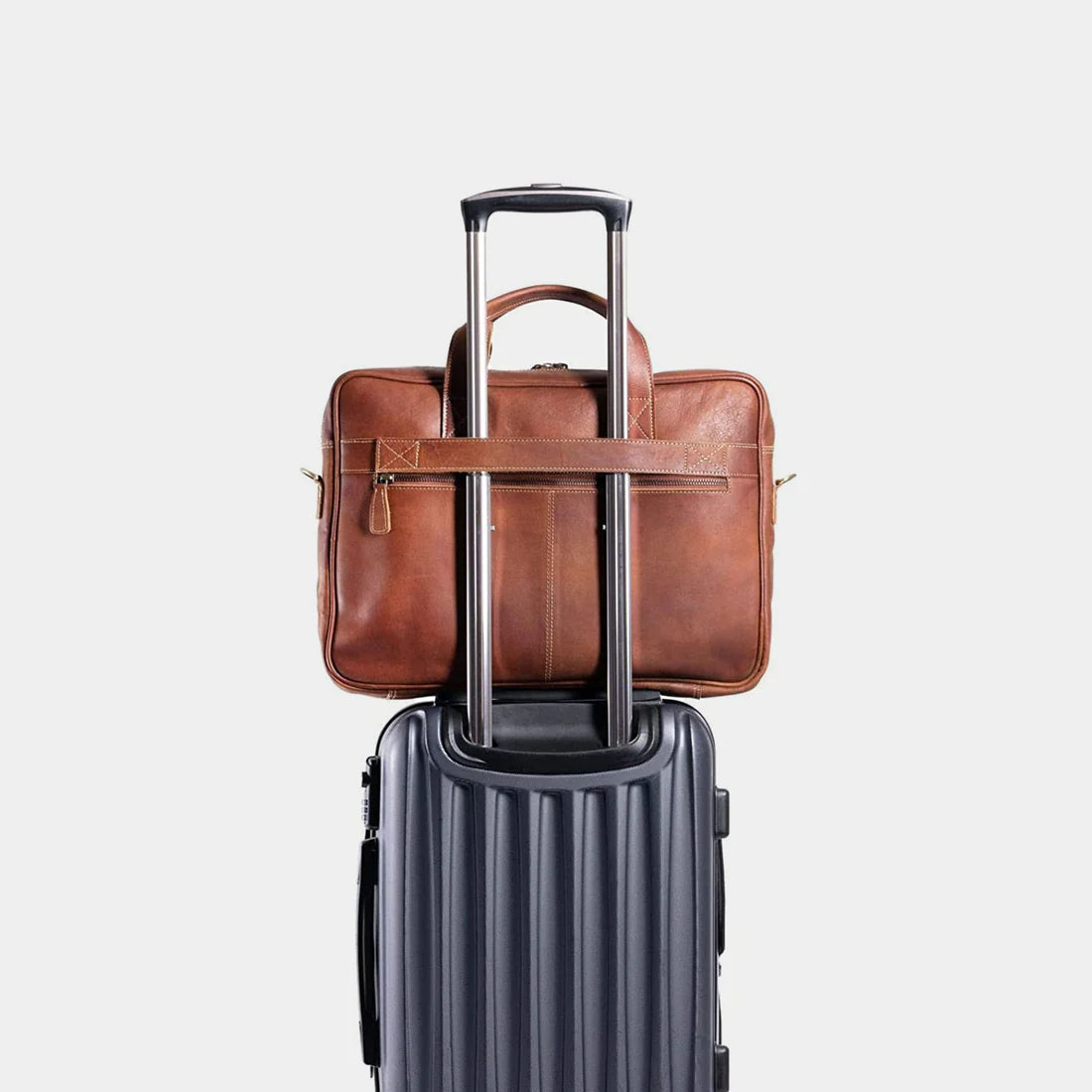 A brown leather briefcase with a horizontal strap across the front, shown stacked on top of a dark gray hardshell rolling suitcase against a light gray background.