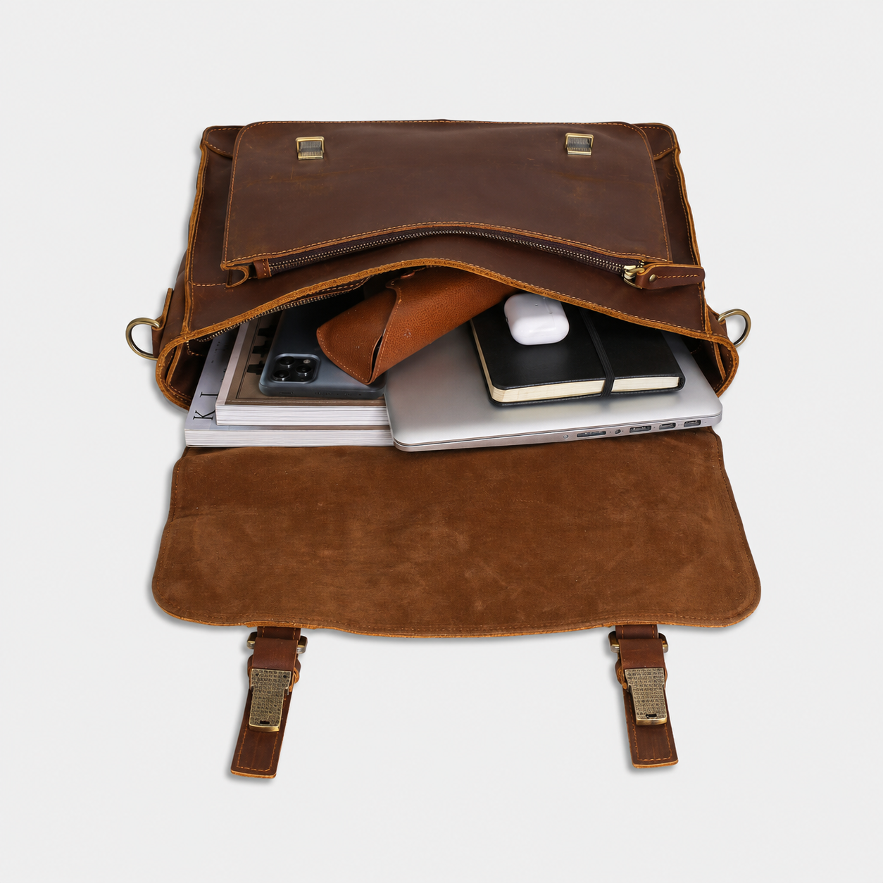 An open brown leather briefcase or laptop bag shown from above on a light gray background, with a laptop, phone, notebook, and earbud case partially tucked inside.